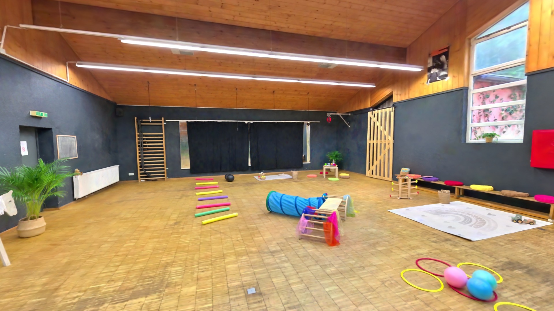 Kameraflug durch die Spiel- und Bewegungsbereiche der Kinderspielgruppe, vorbei an Spielmatten, Reifen, Tunnel und Balancierparcours.